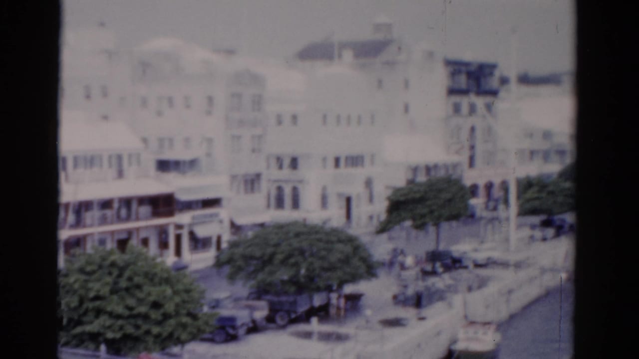 1975 Florida waterfront view with white multi-story buildings, balconies, a tree-lined street, parked cars, and distant people near the seawall.