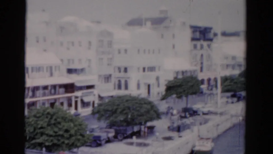 1975 Florida waterfront view with white multi-story buildings, balconies, a tree-lined street, parked cars, and distant people near the seawall.