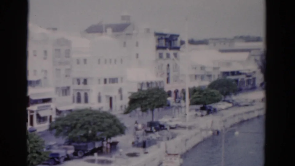 1975 Florida 8mm frame showing white waterfront buildings, a tower-like facade, trees, cars, pedestrians, and a wider stretch of water.