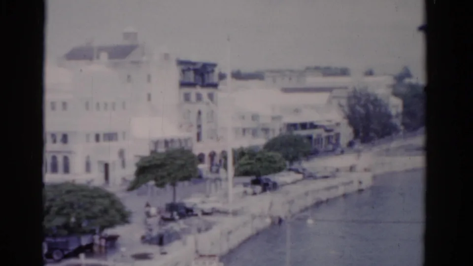 1975 Florida waterfront frame with white buildings, trees, cars, small pedestrians, open water, and a distant large vessel near the horizon.