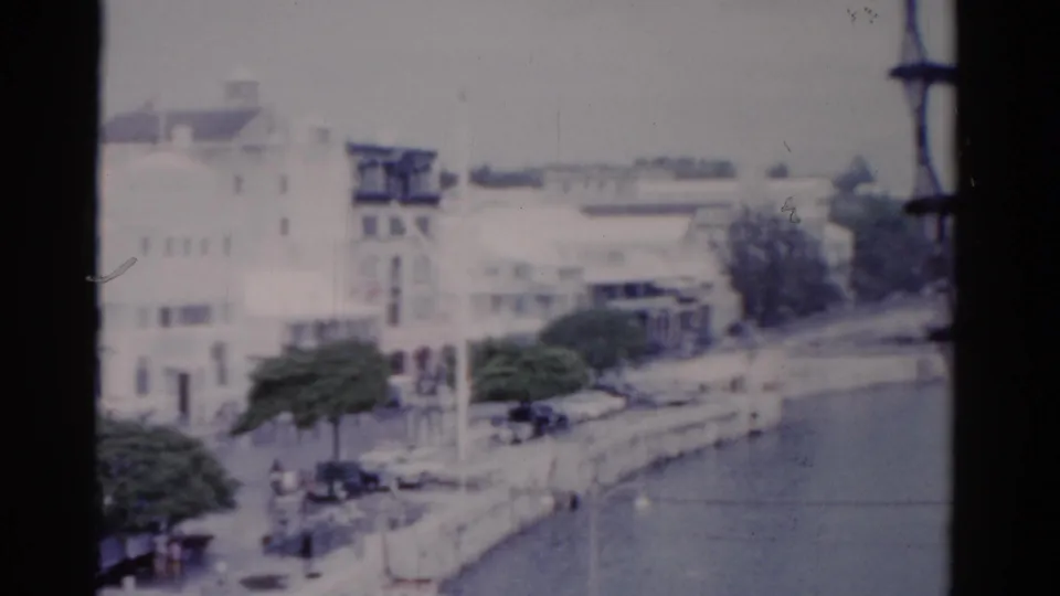 1975 Florida 8mm harbor view showing white waterfront buildings, trees, parked cars, and calm water from an elevated vantage.