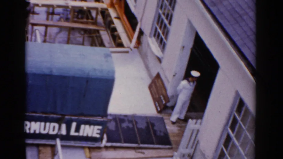 Angled 1975 Florida 8mm view of the Bermuda Line entrance, blue awning, building windows, and a sailor in white at the doorway.