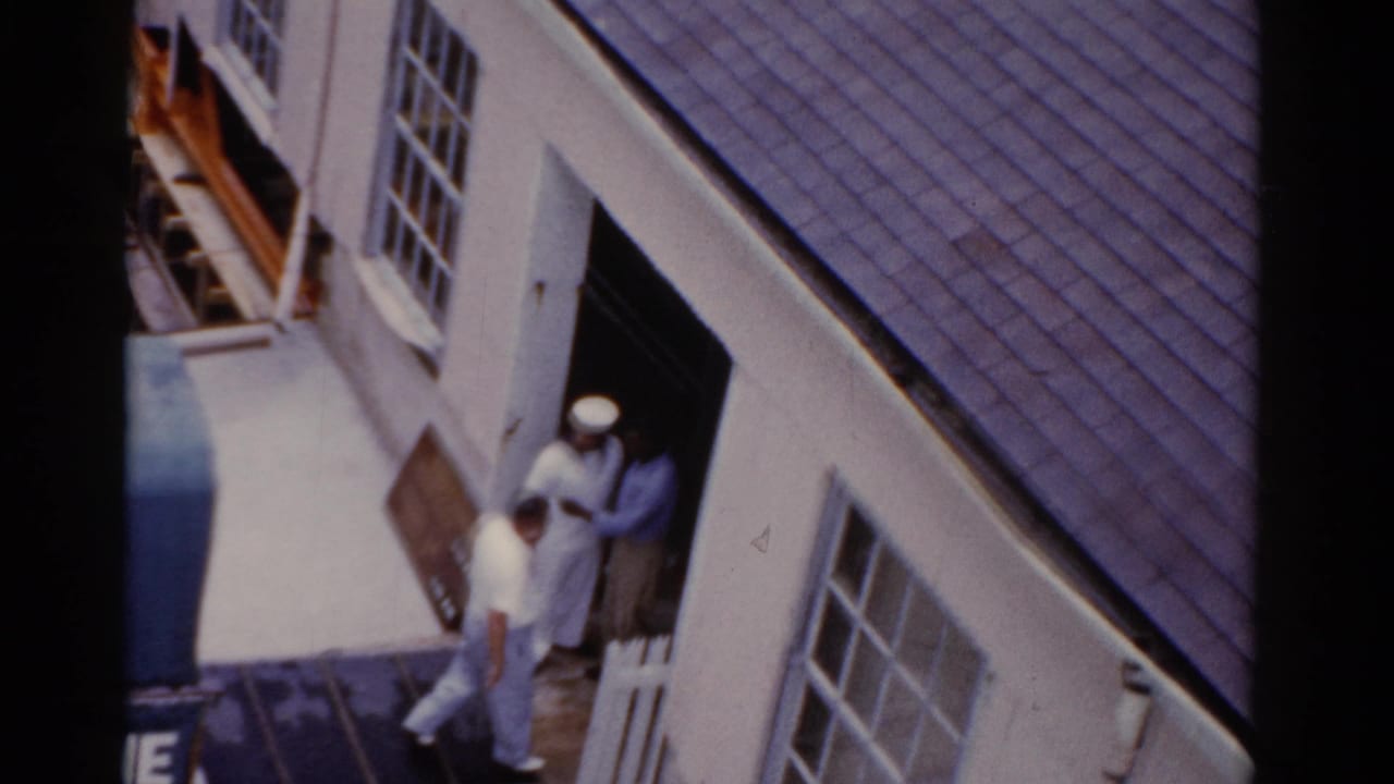 Man in light clothing walks past a sailor by a building doorway.