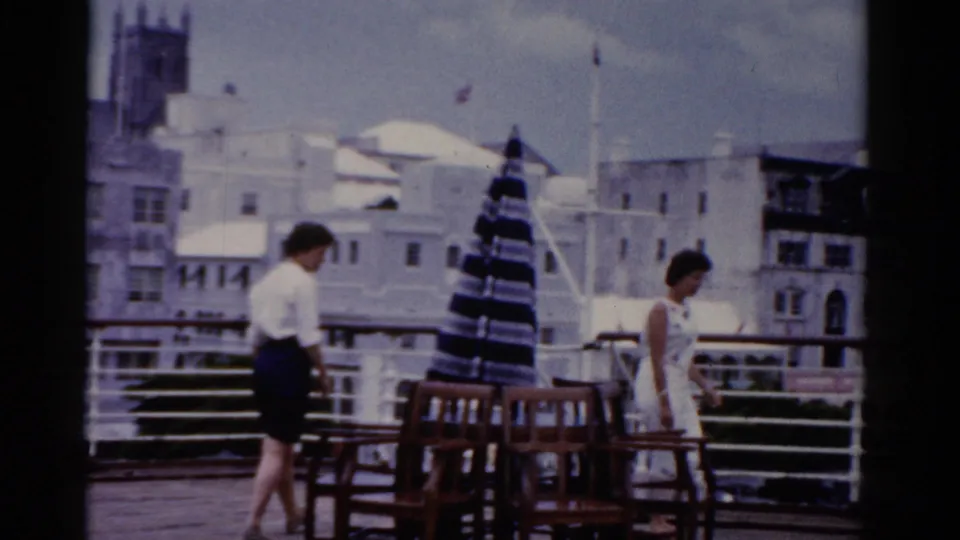 1975 Florida 8mm frame showing adults in summer clothing on a waterfront terrace with chairs, umbrellas, and buildings beyond.