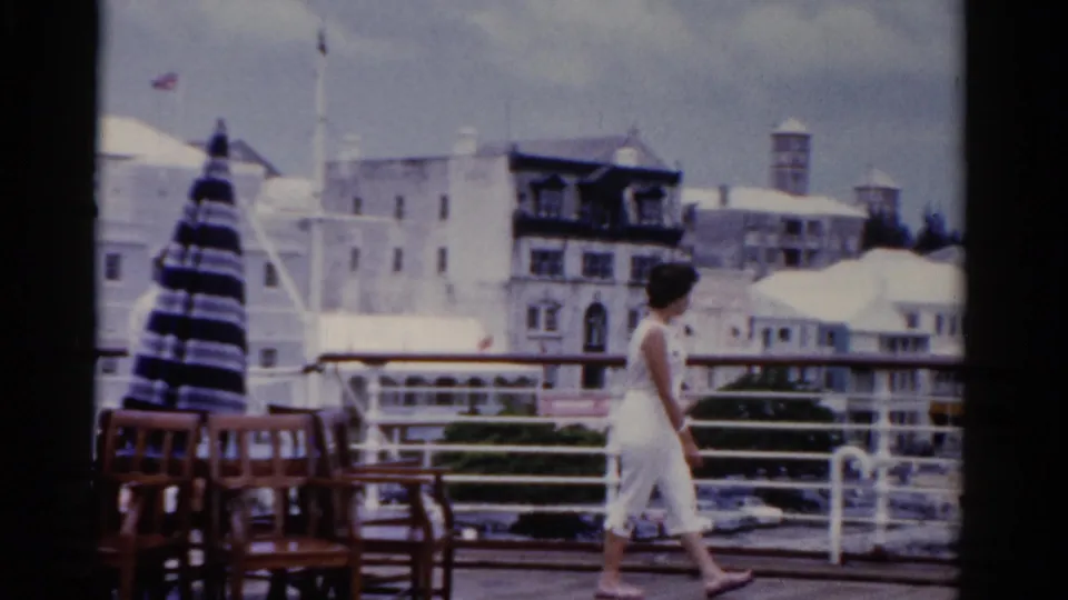 1975 Florida home-movie frame of a woman walking along a waterfront terrace with patio chairs and town buildings behind her.