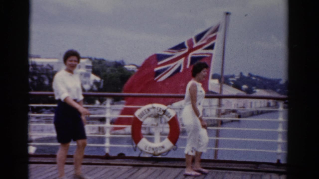 1975 Florida 8mm frame showing two adult women on a ship deck with white railings, water, shoreline, a red life ring, and a large Union Jack-style flag.