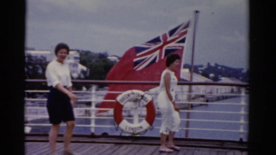 1975 Florida 8mm frame showing two adult women on a ship deck with white railings, water, shoreline, a red life ring, and a large Union Jack-style flag.