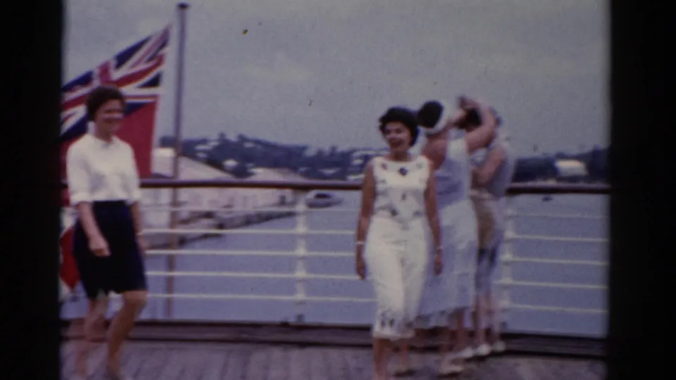 1975 Florida 8mm frame showing a woman in a white patterned outfit walking forward on a ship deck, with other women, railings, water, and flag behind her.
