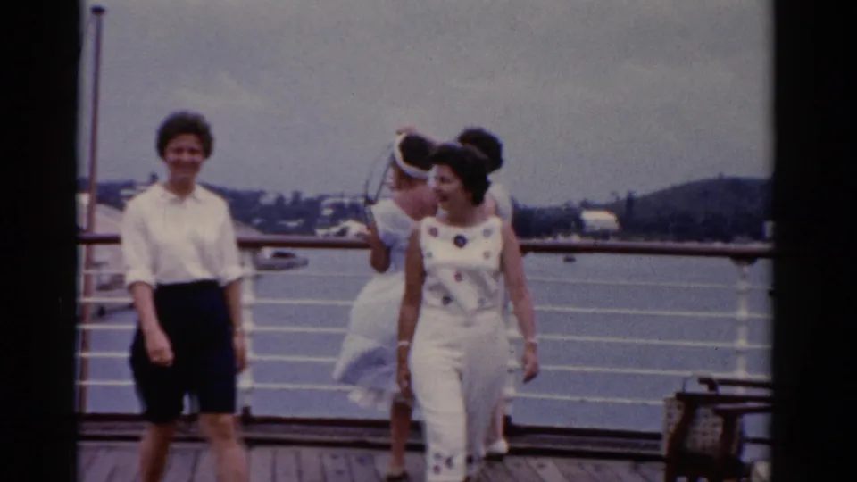 1975 Florida home-movie frame of two adult women walking across a ship deck, with other passengers by the rail and water and shoreline in the background.
