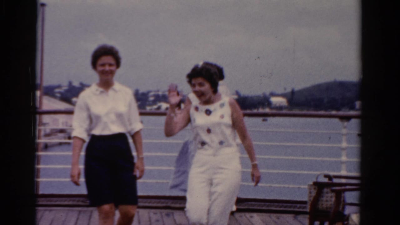 Two adult women in 1970s clothing walk toward the camera on a Florida waterfront deck, with railings and buildings in the background.