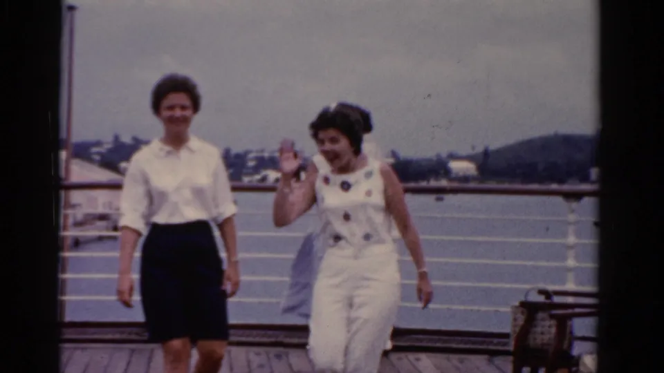 Two adult women in 1970s clothing walk toward the camera on a Florida waterfront deck, with railings and buildings in the background.