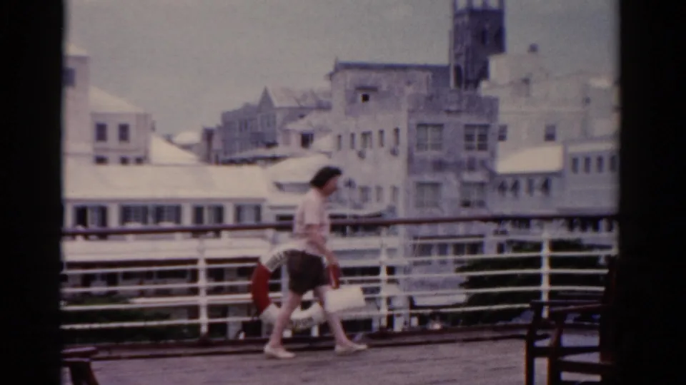 An adult woman walks across a waterfront ship or dock deck, with white railings, a red life preserver, and coastal buildings behind her.