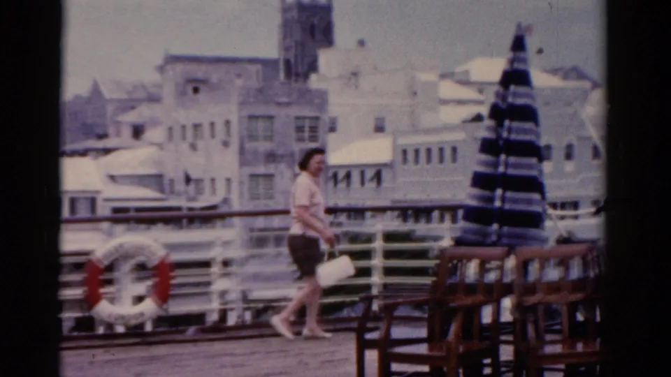 An adult woman walks along the ship or dock deck near railings, a life preserver, benches, and a striped umbrella in 1975 Florida footage.