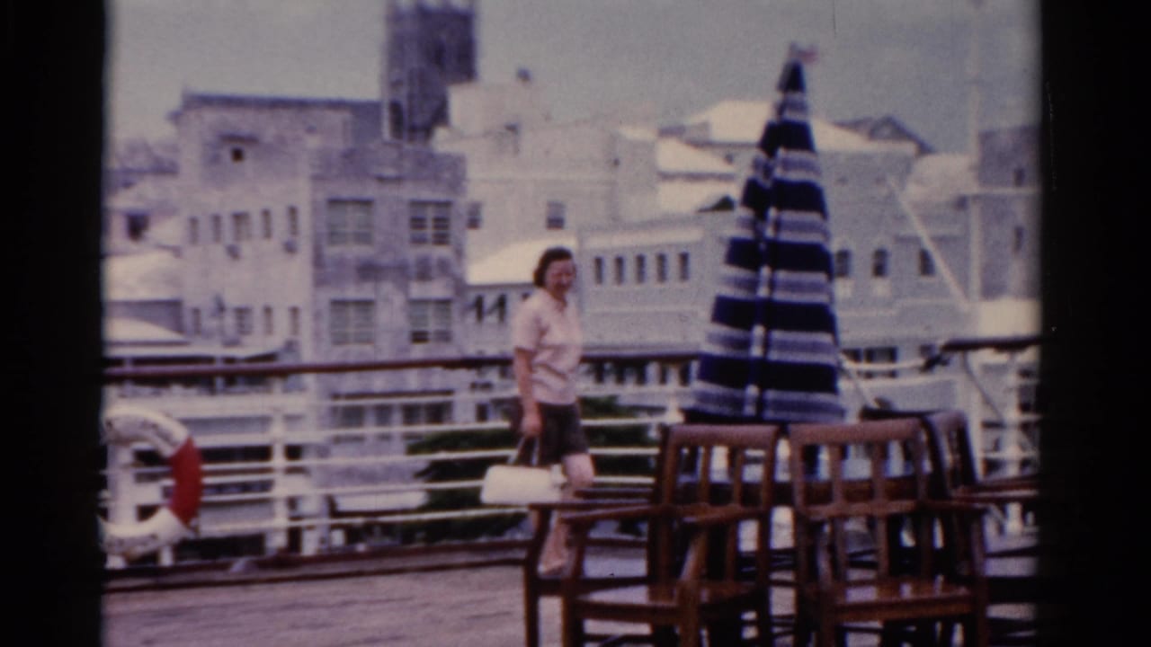 In 1975 Florida home-movie footage, a woman carrying a white handbag walks on an outdoor deck near chairs and a closed striped umbrella.