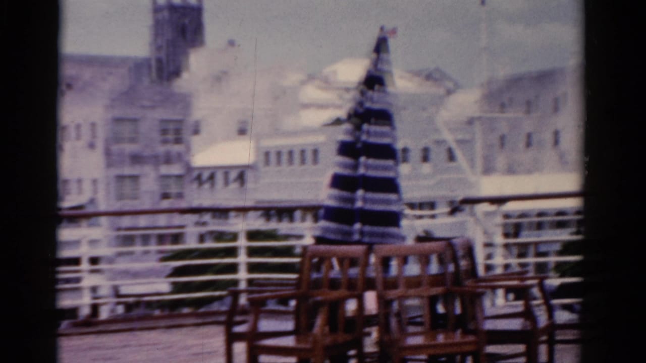 The woman has moved out of frame as the camera holds on deck chairs, a closed striped umbrella, railings, and white waterfront buildings.