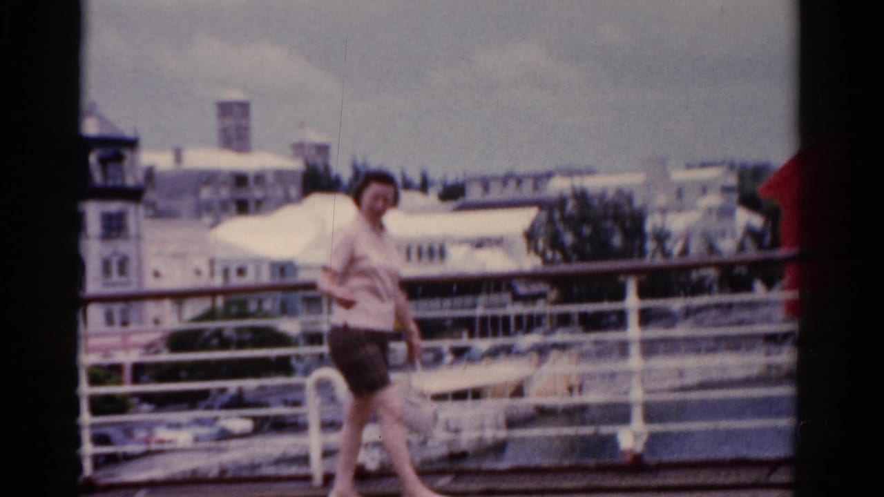 Woman walking on a ship deck with white railings and waterfront buildings behind her.
