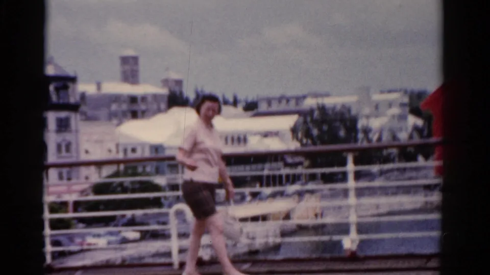 In 1975 Florida footage, a woman in a light blouse and dark shorts walks along a ship deck with harbor buildings in the background.
