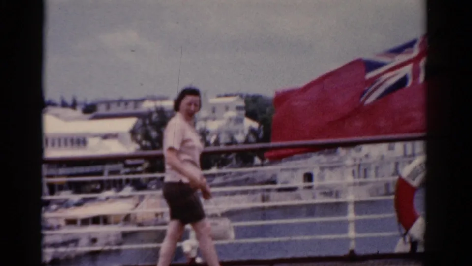 The woman continues along the deck as a large red British flag enters the frame beside white ship railings.