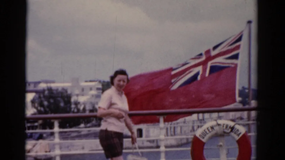 A 1970s home-movie frame shows the woman beside a large red British flag on a ship deck, with harbor scenery behind her.