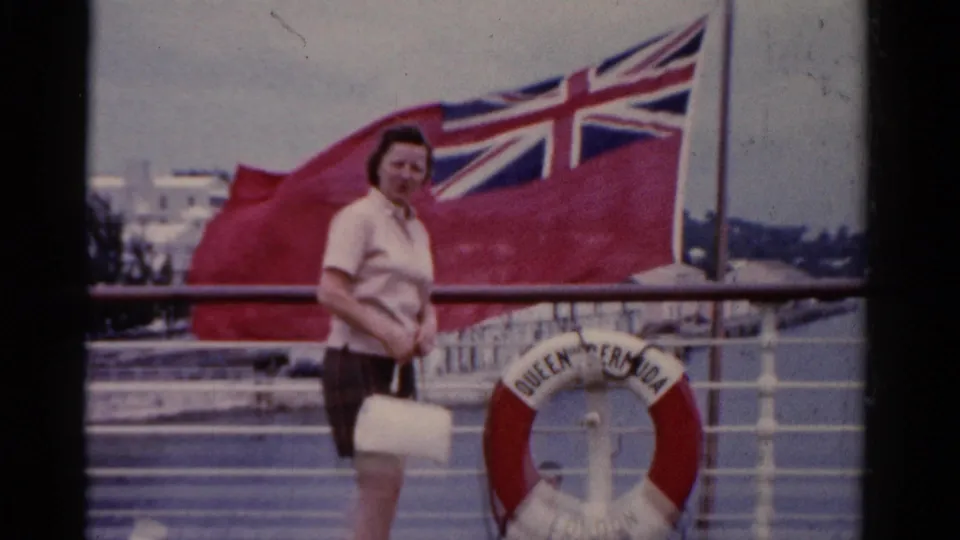 The woman pauses or slows near a life preserver marked with vessel text, with a red British flag and harbor backdrop visible.