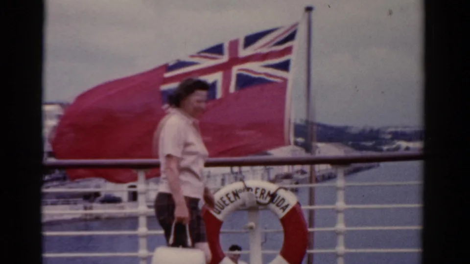 In 1975 Florida footage, a woman walks on a ship deck beside white railings, a British Red Ensign, and a QUEEN OF BERMUDA life preserver.