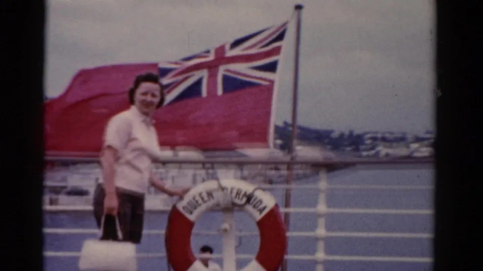 The 1975 home-movie frame shows a woman posed on a ship deck with a white bag, British Red Ensign, and QUEEN OF BERMUDA life preserver.