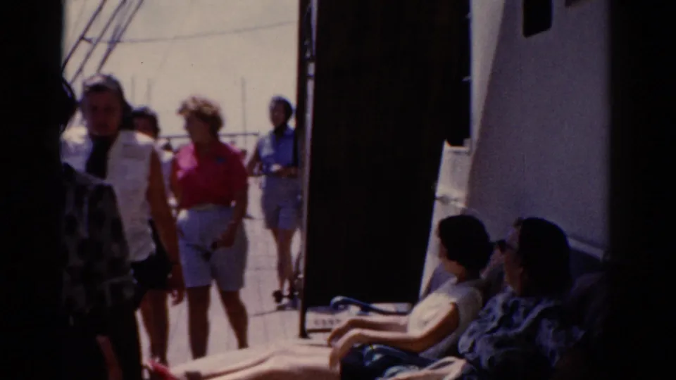Passengers on a 1975 ship deck in Florida walk through a narrow deck area while others sit in the shade along the side of the vessel.