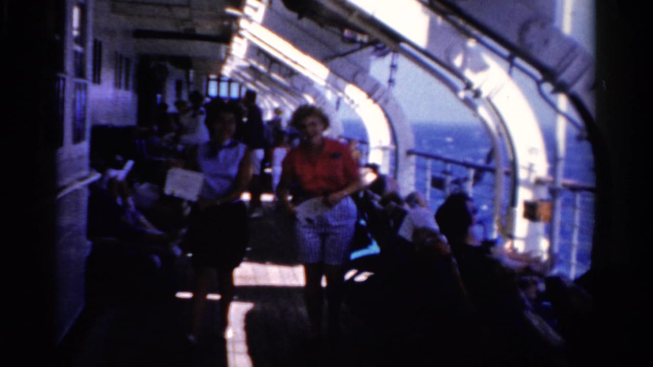 Two women walk along a sunlit cruise ship deck corridor with seated passengers nearby.