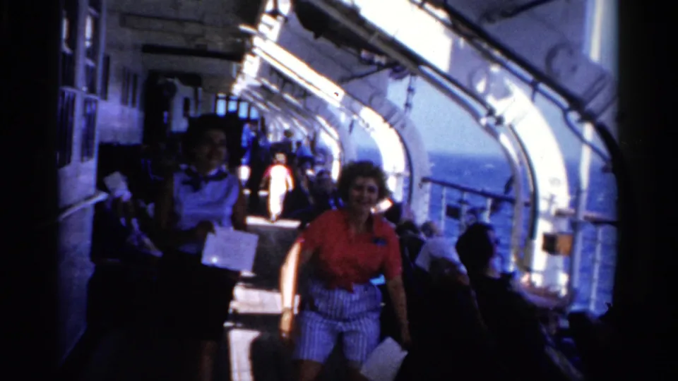 Two smiling women move closer on a 1975 Florida cruise ship deck, with seated passengers and blue sea visible behind them.