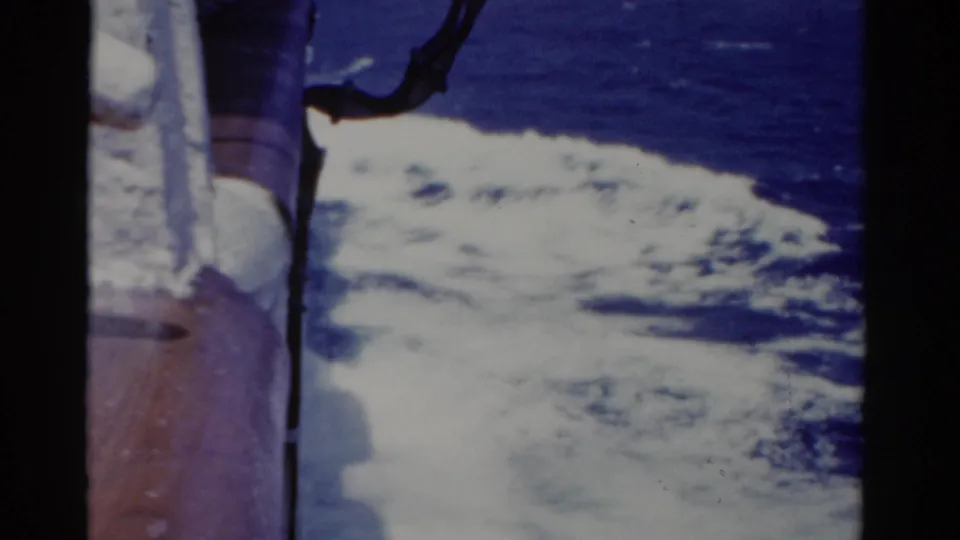 A handheld 1975 cruise-ship view shows foamy wake alongside the vessel, with blue ocean and deck hardware at frame edge.