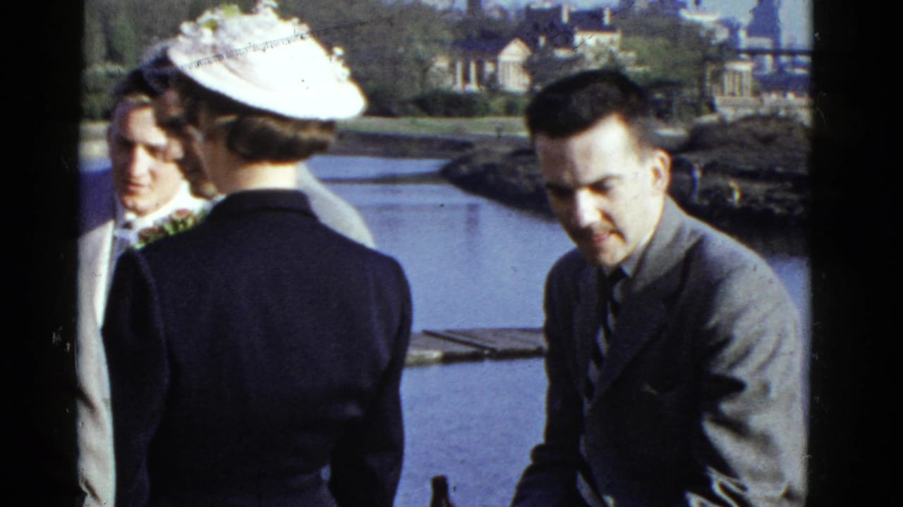 1975 Boston 8mm frame showing adults in suits and a woman in a white hat gathered near water with houses in the background.