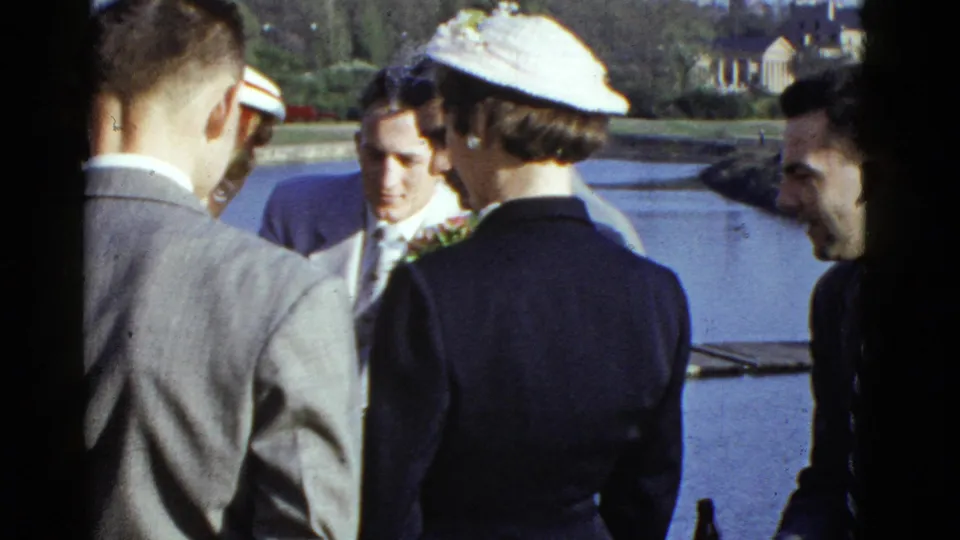 1975 Boston 8mm frame showing a small formal group near water, with men in suits and women wearing hats.