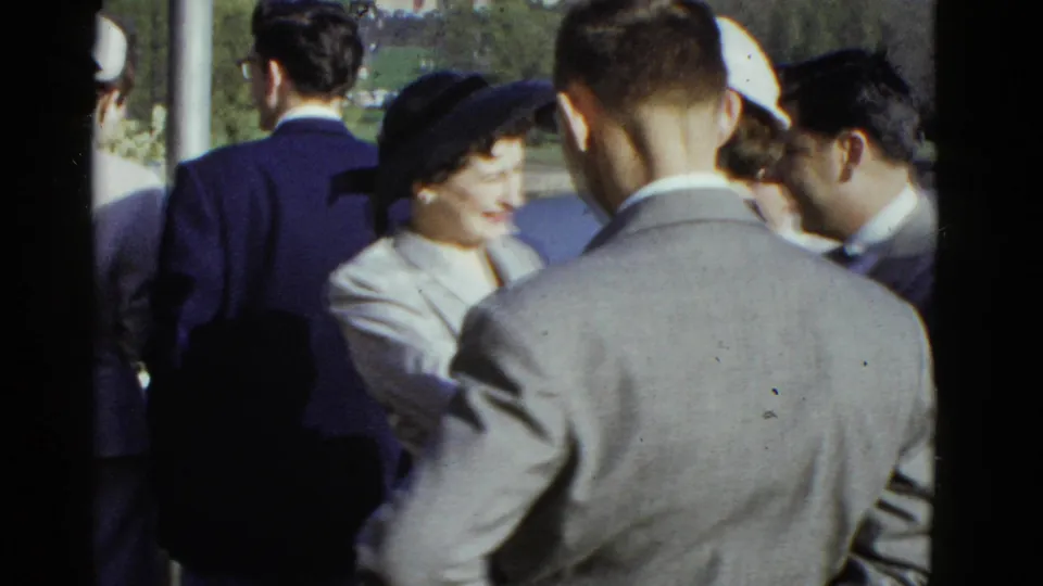 1975 Boston home-movie frame with a woman in a dark hat smiling while adults in suits gather around her outdoors.