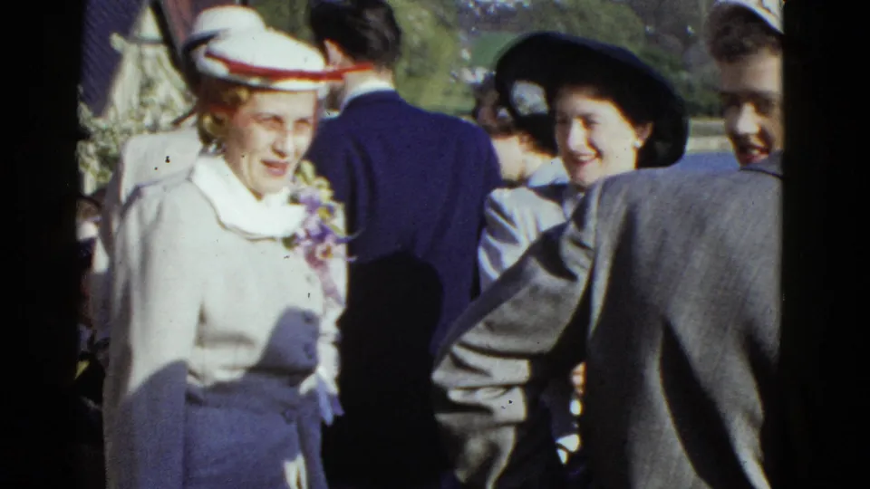In supported 1975 Boston home-movie footage, women in hats and tailored outfits stand close together at an outdoor formal gathering.