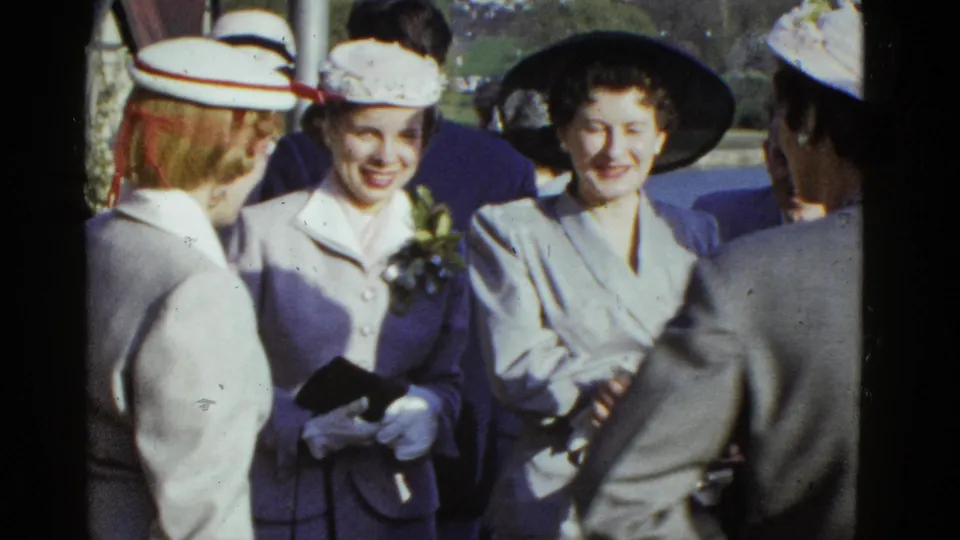 Women at a Boston outdoor gathering smile toward the camera in 1970s formal hats, gloves and tailored clothing.