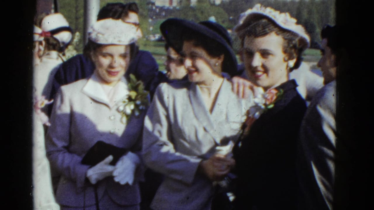 1975 Boston 8mm frame showing women in vintage hats and tailored suits smiling, with one arm around another at an outdoor gathering.