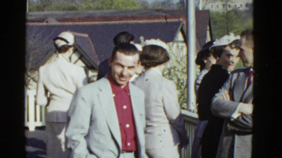 The man remains centered and smiling in the 1975 Boston scene, with women in hats and suited adults visible near a railing and building roofline.