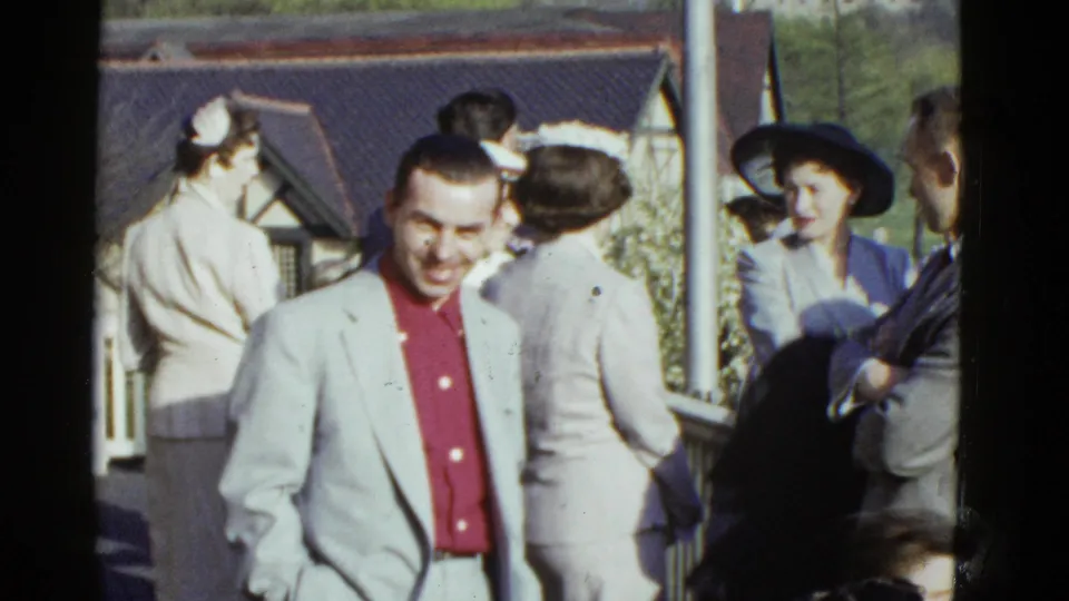 A candid 1975 Boston frame shows the smiling man in a pale jacket foregrounded as adults in hats and spring clothing gather behind him.
