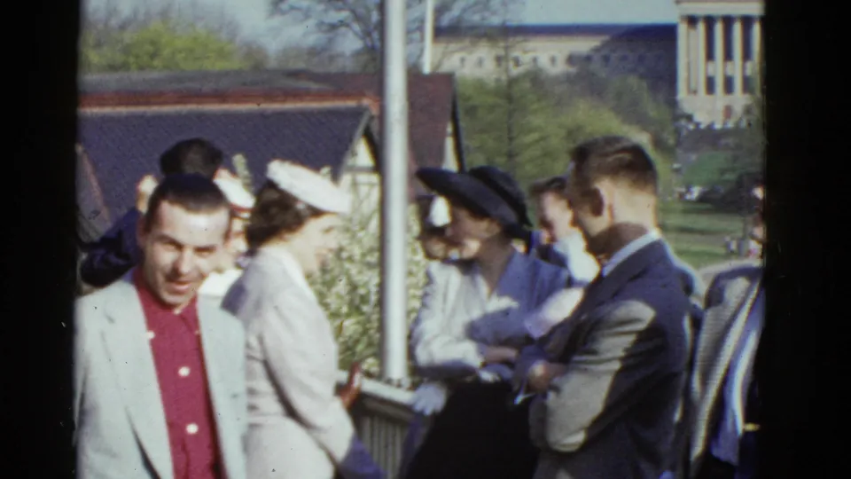 The final frame widens the outdoor Boston gathering, with the smiling man at left and adults in hats and suits by the railing and greenery.