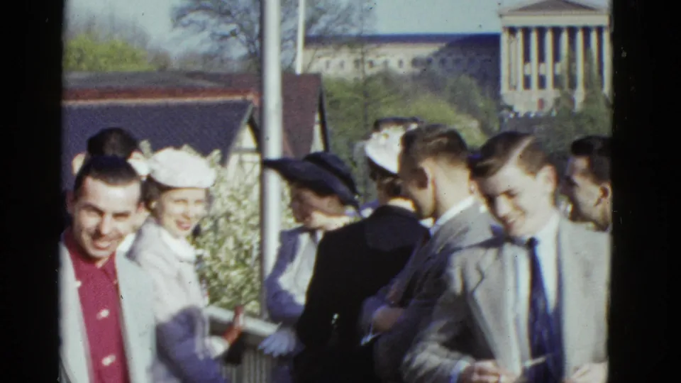 1975 Boston 8mm frame of well-dressed adults smiling and talking outdoors near a distant columned building.