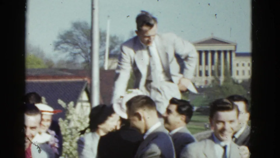 1975 Boston home-movie frame showing a crowded formal outdoor group as one man stands higher than the others.