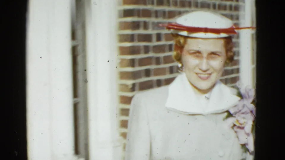 Close 1975 Boston 8mm frame of a woman in a white hat and pale coat smiling near a brick exterior wall.