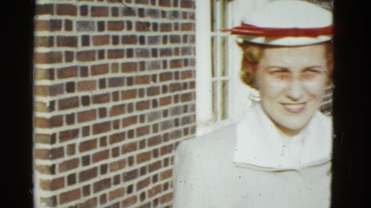 Adult woman in formal dress and a white hat with red trim stands near a brick facade in 1975 Boston home-movie footage.