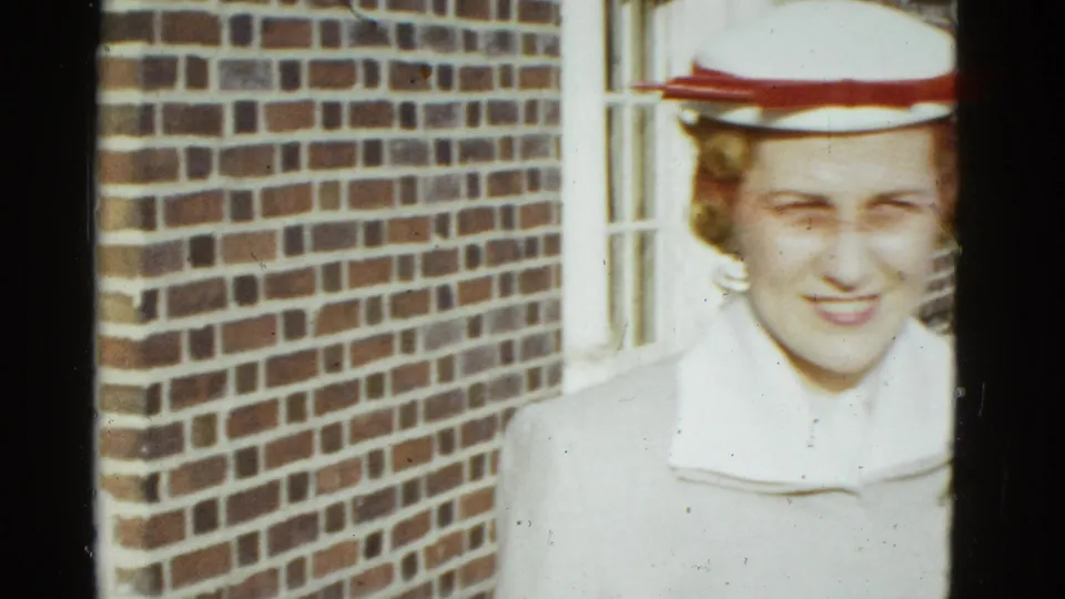 Adult woman in formal dress and a white hat with red trim stands near a brick facade in 1975 Boston home-movie footage.