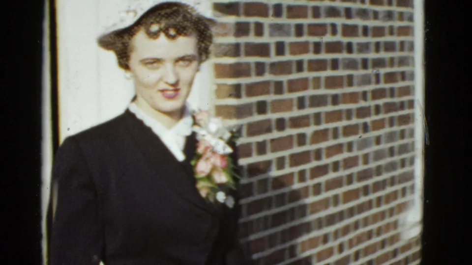 Adult woman in a dark formal suit and small hat faces the camera outside a brick building, wearing a floral corsage.
