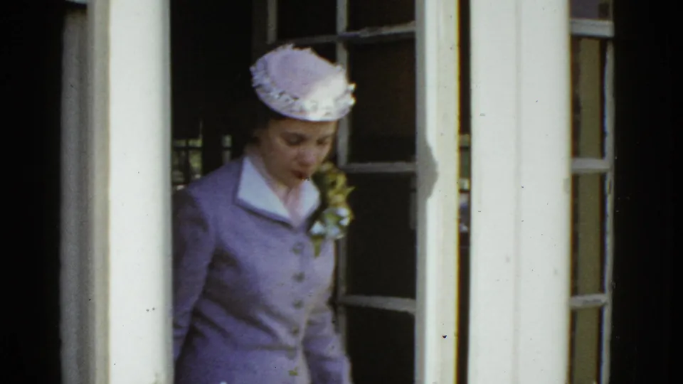 Adult woman in a purple suit and matching hat exits a doorway with a corsage in 1975 Boston home-movie footage.