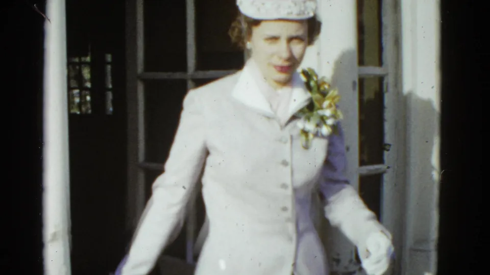 In supported 1975 Boston home-movie footage, a woman in a light suit and matching hat steps from a doorway on a brick porch.