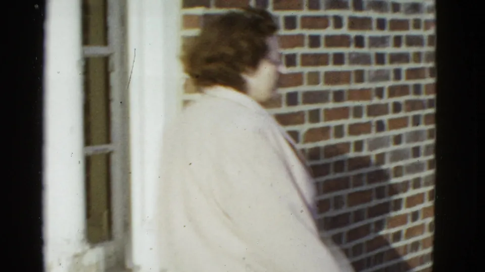 Seen from behind, the woman stands near the brick wall and doorway, with her light jacket and short dark hair visible.