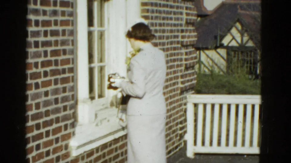 On the brick porch, the woman in a light suit looks down at something she is holding, with windows and railing behind her.