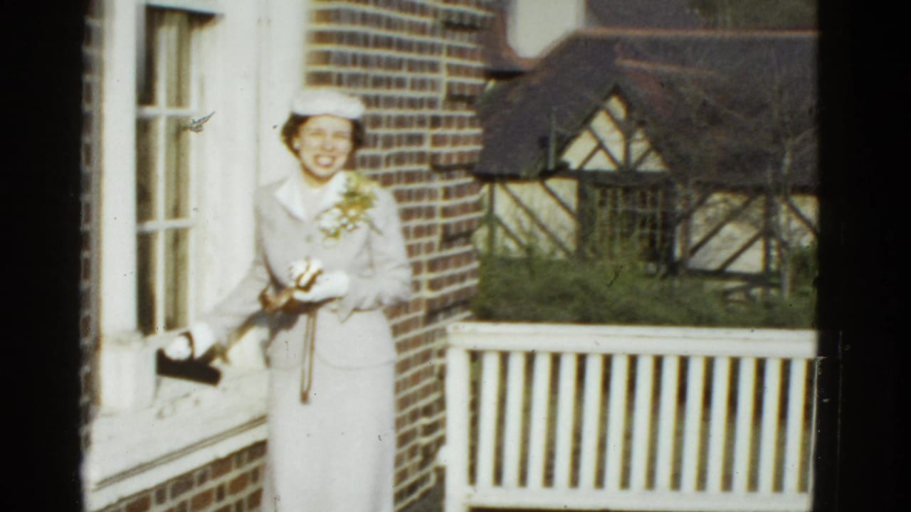 A smiling woman in a light suit and hat holds a camera beside a white porch railing in 1975 Boston footage.